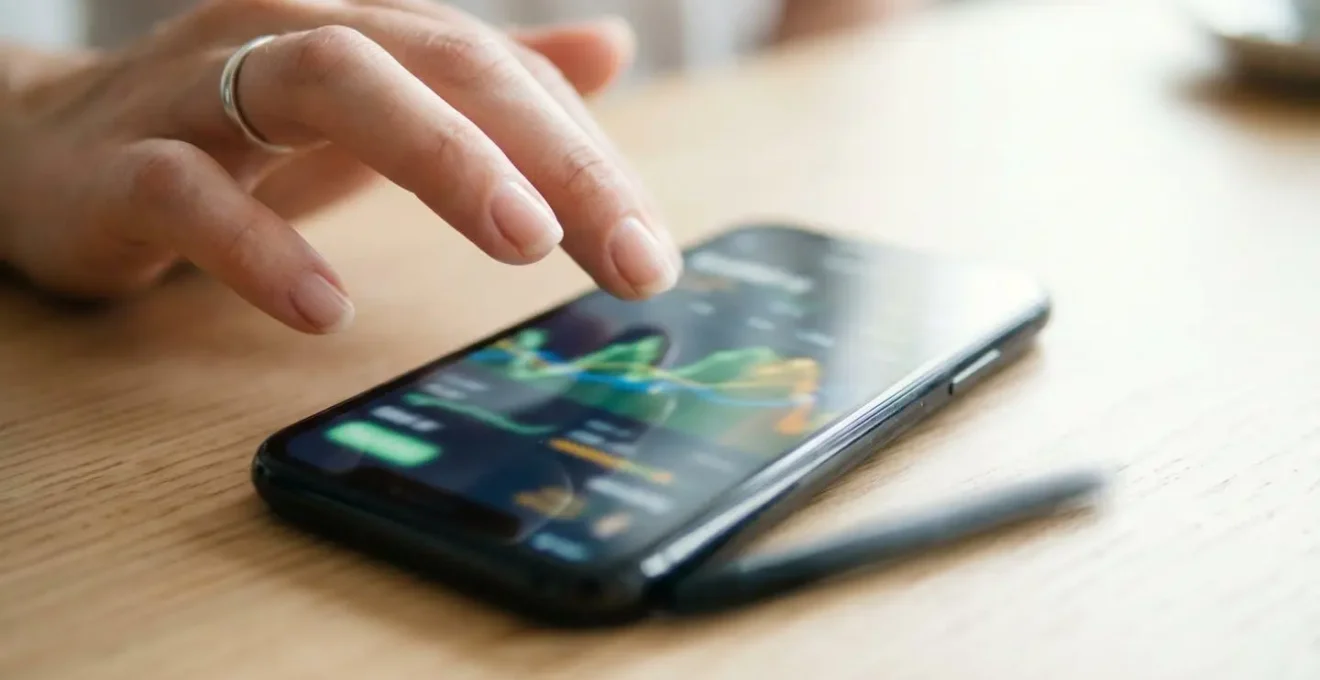 Primo piano di una mano che scorre con il dito su uno smartphone mostrando un grafico di andamento prezzi con schermo parzialmente sfocato, su superficie di legno chiaro con sfondo luminoso bokeh